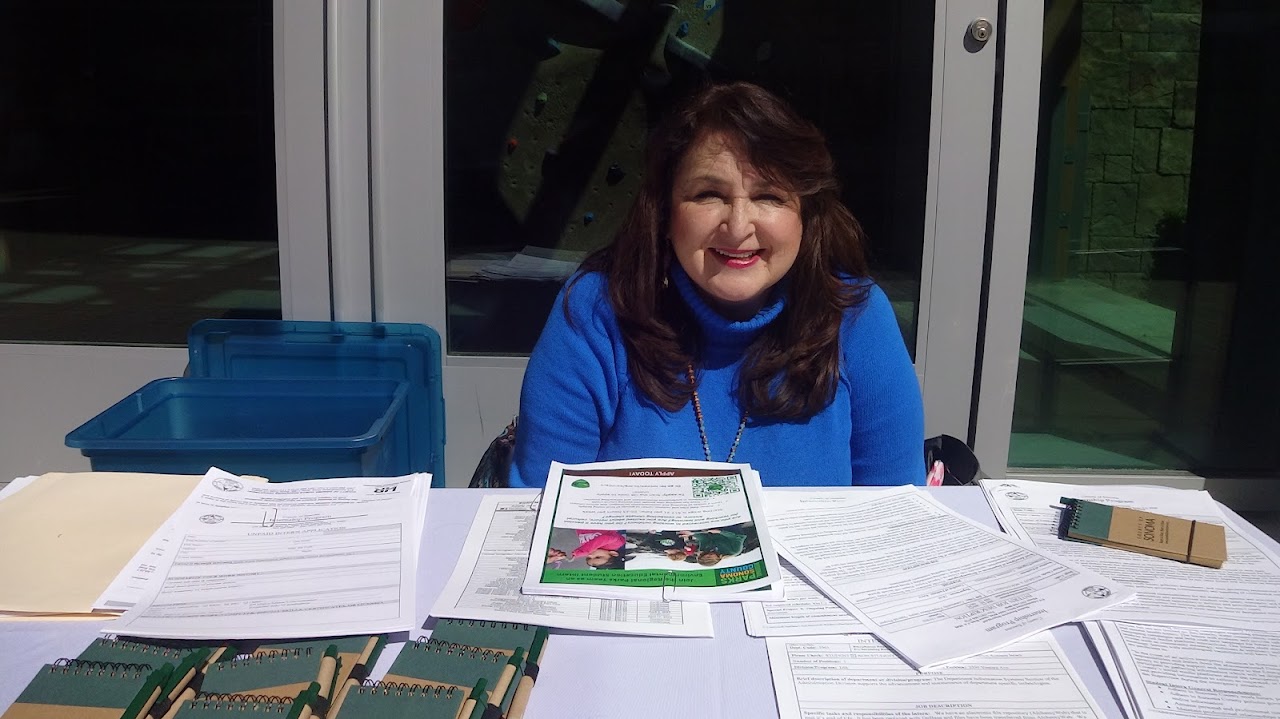 Adriana Call sitting down outside in front of her table smiling at the camera. There are recruitment materials on the table. 