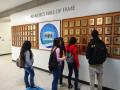 4 students are standing in front of a wall of sports plaques