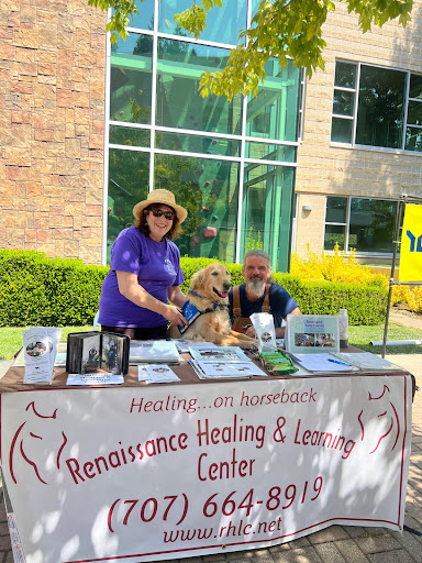 A woman in a purple shirt stands behind a table posing next to a dog in a harness. Beside them is a man in brown overalls smiling at the camera. 